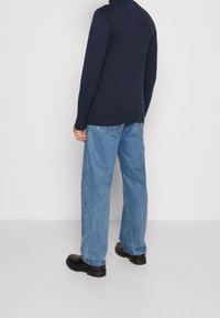 Persona che indossa una maglia a maniche lunghe blu scuro, jeans azzurri e scarpe nere robuste, in piedi davanti a uno sfondo bianco uniforme.