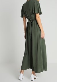 Dlouhé olivově zelené šaty s volnými rukávy, páskem v pase a splývavou sukní. Nosené s bílými botami na blokovém podpatku.