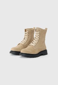Botas de tobillo de ante beige con cordones en la parte delantera, cremallera lateral y suela de goma negra que presenta patrones texturizados y puntera redonda.