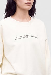 Femme portant un sweat-shirt couleur crème avec le nom de la marque en relief "MICHAEL KORS" sur la poitrine, coupé au niveau du cou et des épaules.