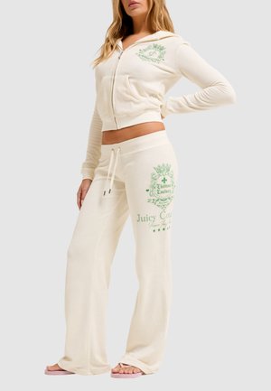 Vrouw draagt een crèmekleurige Juicy Couture velours hoodie en bijpassende broek met groene logo-opdruk, gecombineerd met roze slippers.