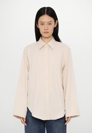 Lys beige langermet bluse med spiss krage, knappfeste foran og løs passform. Stoffet ser glatt ut med en svak glans.