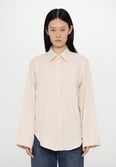 Blusa a maniche lunghe color beige chiaro con colletto a punta, chiusura frontale a bottoni e vestibilità ampia. Il tessuto appare liscio con una leggera lucentezza.