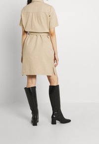 Femme portant une robe beige à manches courtes ceinturée et des bottes en cuir texturé noires montantes jusqu'aux genoux, debout devant un fond blanc uni.