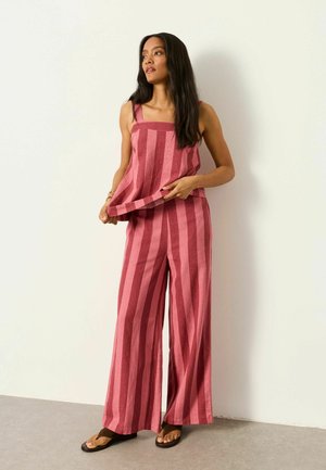 Frau trägt pink-rot gestreiftes ärmelloses Top und weit geschnittene Hosen, steht vor einer schlichten weißen Wand und hält den Saum des Tops.