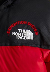 Czerwona i czarna kurtka o teksturowanym wykończeniu. Posiada haftowane logo "SYSTEM EKSPEDYCJI" oraz "THE NORTH FACE" w białym i czerwonym kolorze.