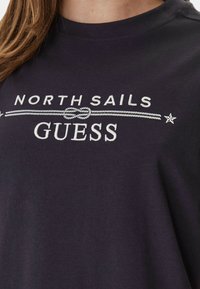 Sweatshirt foncé avec les textes "NORTH SAILS" et "GUESS", séparés par un graphique de corde et de nœud, porté par une personne aux cheveux bruns.