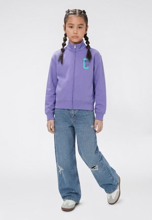 Junges Mädchen mit geflochtenen Haaren, das eine lila Reißverschlussjacke, weit geschnittene zerrissene Jeans und weiße Turnschuhe trägt und vor einem weißen Hintergrund steht.