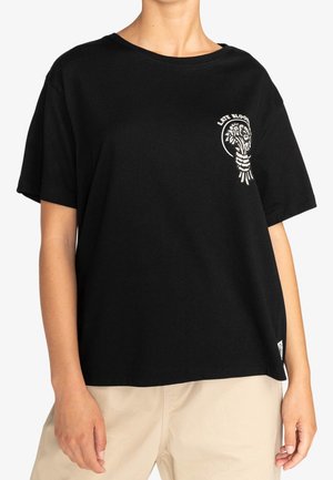 Czarny T-shirt z krótkim rękawem z małym białym kwiatowym wzorem i napisem "LATE BLOOM" na górnej lewej części klatki piersiowej, noszony z beżowymi spodniami.