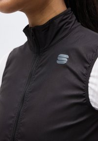 Inimese lähipilt, kes kannab valge pika varrukaga särgi peal musta varrukateta lukuga vest, mille rinnal on väike peegelduv logo.