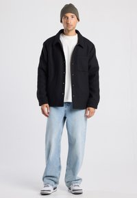 Black button-up jacket with a collar and chest pocket, paired with light blue jeans and a brown knit beanie. Casual footwear completes the look.