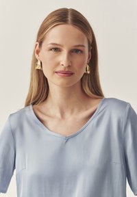 Hellblaue Kurzarmbluse mit V-Ausschnitt, die eine glatte Textur und dezente Steppnähte aufweist. Goldene Statement-Ohringe runden den Look ab.