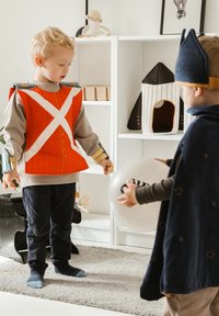 Twee kinderen in kostuums: de ene draagt een rood vest met witte strepen en blauwe accenten, de ander een donkere cape en houdt een doorzichtige ballon vast.