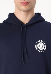 Felpa blu navy con tasca a canguro, cappuccio con coulisse e logo bianco Lacoste Paris con design di tennis. Tessuto morbido.