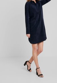 Mörkblå långärmad klänning i mjukt tyg, med krage, sned design och rundad nederkant, ihopparad med svarta klackar med ankelrem.