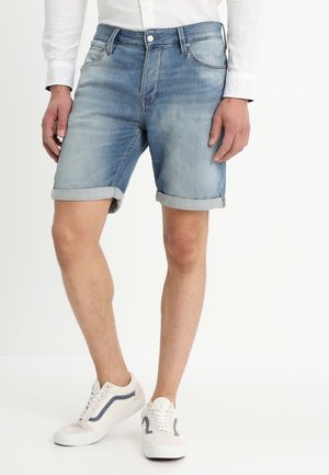 Pantalones cortos de mezclilla azul claro con dobladillos enrollados, cinco bolsillos y cierre de botón. Combinados con zapatillas blancas con acentos azules.