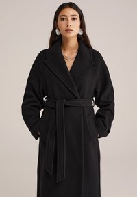 Schwarzer Wickelmantel aus weichem Material, mit breitem Kragen, langen Ärmeln und einem gebundenen Gürtel. Enthält seitliche Taschen für zusätzliche Funktionalität.