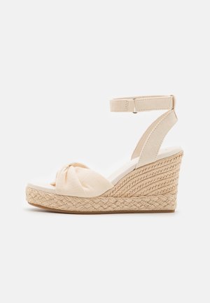 Beige wedge sandaal met een geweven jute zool, bovenwerk van zacht stof met knoopdetail en een stoffen enkelband voor een veilige pasvorm.