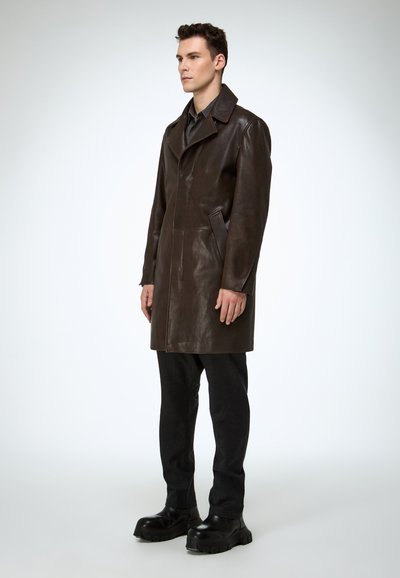 Brauner Ledermantel mit einem Reverskragen, Knopfverschluss, seitlichen Taschen und einer schlanken Silhouette, kombiniert mit dunkler Hose und klobigen schwarzen Schuhen.