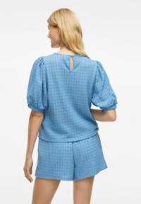 Top azzurro chiaro con texture, maniche a sbuffo e un piccolo fori sul retro, abbinato a pantaloni corti coordinati. Entrambi i pezzi presentano un motivo tessuto.