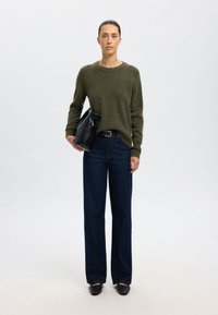 Pulover verde măsliniu, blugi largi de culoare albastru închis, geantă neagră cu mâner curbat și pantofi negri sport. Design simplu și curat, cu hardware minim.