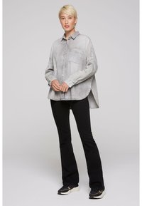 Gray oversized button-up shirt with a single chest pocket, long sleeves, and curved hem, paired with black flared pants and sneakers.