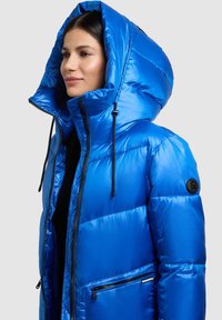 Pūsta mėlyna striukė su blizgiu paviršiumi, aukšta gobtuvu, juodomis kišenėmis su užtrauktukais ir reguliuojamais raišteliais. Rankovėje yra logotipo emblema.