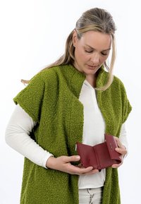 Femme portant un cardigan vert texturé et une chemise blanche, regardant vers le bas tout en tenant un portefeuille en cuir rouge ouvert avec un compartiment zippé.