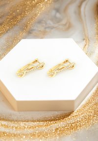 Guldtonade örhängen formade som sammanflätade slingor, med klara rhinestones längs ena sidan och en säker baksida, visade på en hexagonal ställning.