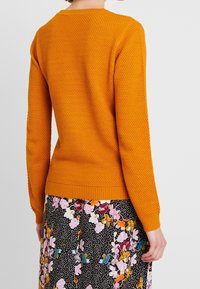 Person som bär en texturerad orange långärmad tröja och en svart kjol med flerfärgade blommor och vita prickar, sedd bakifrån.