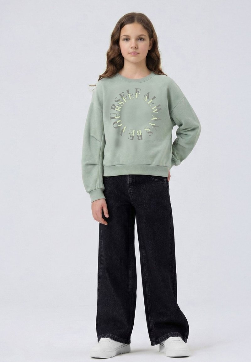 Jeune fille debout, une main sur la hanche, portant un sweat-shirt vert clair avec le texte en cercle "Always Be Yourself", un jean noir à jambes larges, et des baskets blanches.