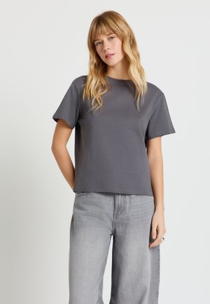 Donna bionda con capelli mossi indossa una t-shirt a maniche corte grigio scuro e jeans a gamba larga grigio chiaro, in piedi davanti a uno sfondo semplice.
