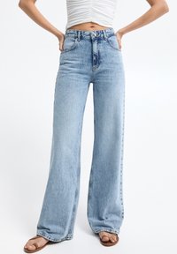 Jean denim bleu clair taille haute à jambes larges, avec fermeture à bouton et zipper, poches avant, et texture légèrement d élavée.