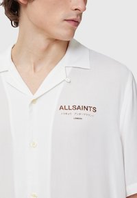 Witte korte mouwen shirt van lichtgewicht stof, met een afgeknipte kraag en "ALLSAINTS" borduursel in bruin op de linker borst.