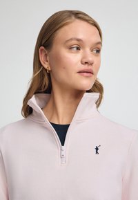 Ljust rosa zip-up sweatshirt med hög krage, svart logotyp på bröstet och slät textur. Under finns en mörk rundhalsad tröja.