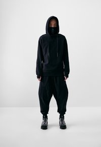 Schwarzer Kapuzenpullover mit Kordelzügen, kombiniert mit weiten schwarzen Hosen mit Taschen. Getragen mit schwarz-grau gemusterten Sneakers.