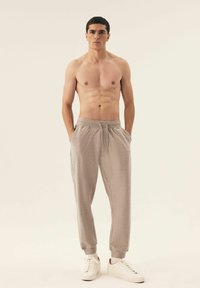 Helle graue Jogginghosen mit elastischem Bund und Bündchen, ausgestattet mit seitlichen Taschen, getragen zu weißen Turnschuhen, Modell steht mit den Händen in den Taschen.