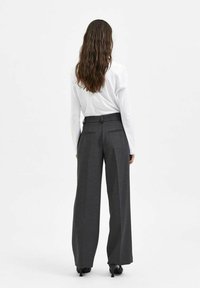 Pantaloni grigi a gamba larga con una vestibilità elegante, dotati di due tasche posteriori e una superficie liscia. Indossati con una camicia bianca a maniche lunghe.