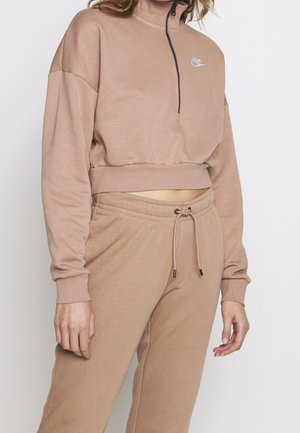 Person iført beige cropped sweatshirt med halvlukning og matchende joggers med snor mod en helt hvid baggrund.