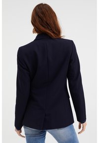 Navy blazer with structured shoulders, slim fit, single vent, and visible stitching details. Worn over light blue jeans.