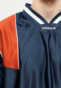Tamsiai mėlynas Adidas sportinis marškinėlis su balta dryžuota apykakle ir oranžiniu tinkleliu peties srityje ant žmogaus viršutinės liemens dalies.