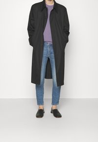 Abrigo largo negro, abotonado, con cuello. Lleva puesto sobre una camiseta púrpura y unos jeans de mezclilla azul claros. Unos zapatos negros tipo slip-on completan el atuendo.