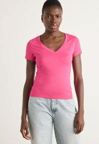 Roze V-hals t-shirt van gladde stof, met korte mouwen en een klein logo op de linkerborst. Draagt met lichtblauwe jeans.