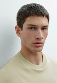Beige mikina s kulatým výstřihem a krátkými rukávy, měkká struktura a minimalistický design. Neutrální výraz modelu doplňuje jednoduchou estetičnost.