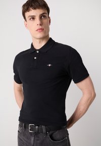 Black polo shirt with short sleeves, two-button collar, and a small embroidered logo on the chest, paired with black jeans and a belt.