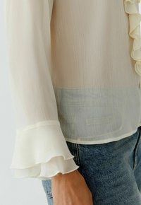 Transparente, leichte cremefarbene Bluse mit Rüschenausschnitt und ausgestellten Ärmeln, kombiniert mit Denim. Der Stoff weist ein dezentes strukturiertes Muster auf.