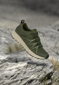 Πράσινο ορειβατικό παπούτσι με χοντρή λευκή σόλα στον αέρα πάνω από βραχώδες έδαφος με μικρά φυτά και θολό φόντο.