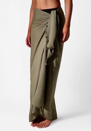 Blootsvoets persoon die een olijfgroene sarong tot aan de enkel draagt, vastgeknoopt in de taille met franjes aan de randen, staand tegen een witte achtergrond.