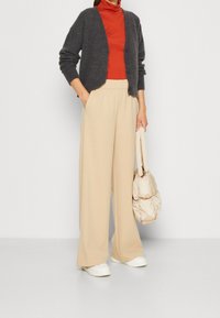 Cardigan grigio con bottoni, maglione rosso a collo alto, pantaloni beige a gamba larga e sneakers color crema. Porta una borsa puffy beige chiaro. Texture liscia.