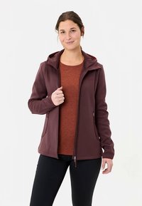 Bordeauxrode fleecejas met capuchon, zakken met ritssluiting en een slanke pasvorm, gedragen over een roestkleurig shirt. Gladde textuur en sportief ontwerp.